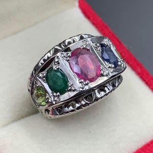Puede incluir: Un anillo de plata con un rubí ovalado, esmeralda y zafiro. El anillo tiene un diseño decorativo alrededor de las piedras preciosas y una piedra verde más pequeña en el lateral. El anillo está en una caja de terciopelo rojo.