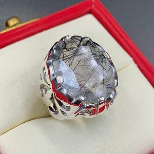 Pode incluir: Um anel de prata com uma pedra preciosa grande e transparente com inclusões pretas. A pedra preciosa, de forma oval, está engastada numa banda de prata decorativa. O anel é apresentado numa caixa vermelha.