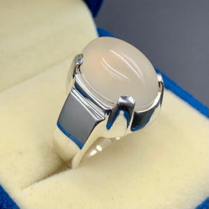 Pode incluir: Um anel de prata com uma grande pedra ovalada esbranquiçada. A pedra é engastada numa banda de prata polida com um design retangular nas laterais. O anel é apresentado numa caixa de joias azul e creme.