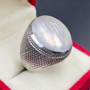 Pode incluir: Um anel de prata com uma grande pedra preciosa ovalada branca leitosa. A banda do anel tem um padrão texturizado de cruzamento. A pedra preciosa é definida em uma moldura lisa e polida. O anel é exibido em uma caixa de joias vermelha.