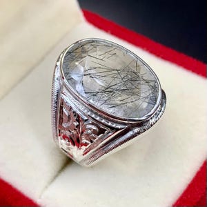 Pode incluir: Anel de prata com uma pedra preciosa oval e transparente com inclusões pretas em forma de agulha. O anel tem um design gravado nas laterais, sobre um fundo de veludo vermelho.