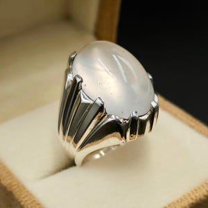 Pode incluir: Um anel de prata com uma grande pedra de lua branca de forma oval. O anel tem um design detalhado e ornamentado.