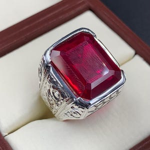Könnte beinhalten: Ein silberner Ring mit einem großen, rechteckigen, tiefroten Edelstein. Der Ring hat ein verziertes, graviertes Design am Band. Der Edelstein ist der Mittelpunkt, fängt das Licht ein und zeigt eine satte, lebendige Farbe.