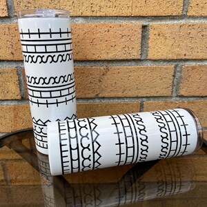 May include: Two white insulated tumblers with black geometric patterns. The design includes horizontal lines, connected X shapes, and rectangular blocks. One tumbler is upright, and the other is lying down, both on a reflective surface.