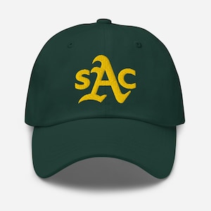 Puede incluir: Gorra de béisbol verde oscuro con visera curva. La gorra presenta las letras bordadas "sAc" en una fuente amarilla estilizada. El sombrero está hecho de una tela suave y tiene una correa ajustable.