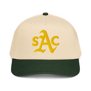 Puede incluir: Gorra de béisbol color crema con visera y botón verde oscuro. La gorra muestra las letras bordadas "SAC" en oro, en una fuente estilizada.