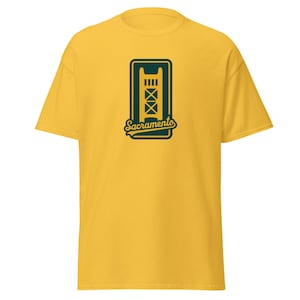 May include: A yellow t-shirt featuring a green and gold graphic of a bridge with the word "Sacramento" in a script font. The t-shirt is made of a soft material and has short sleeves.
