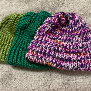 May include: Three knitted beanies in shades of green and purple. The first beanie is a solid light green, the second is a darker green, and the third is a mix of purple, pink, and white yarn with flecks of orange.