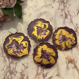 May include: Four purple and gold resin coasters with a marbled design. Each coaster features a gold flower-like pattern.