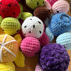 May include: A selection of brightly coloured, crocheted fruit toys. The image includes a red watermelon, green kiwi, pink strawberry, and a purple blackberry. The toys are made of soft yarn and have details like seeds and leaves.