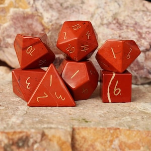 May include: A set of red gemstone dice with gold numerals. The dice are in various geometric shapes, including a d20, d10, d6, and a d4. The dice are displayed on a textured, light brown surface.
