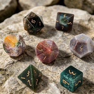 Könnte beinhalten: Sieben Edelsteinwürfel in verschiedenen Formen, darunter d4, d6, d8, d10, d12 und d20. Die Würfel haben Farben wie Grün, Braun, Rot und Grau, mit goldenen Zahlen. Die Würfel liegen auf einer strukturierten Steinoberfläche.