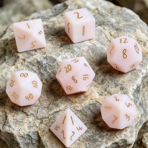 May include: A set of seven pale pink polyhedral dice with gold numerals, including d4, d6, d8, d10, d12, and d20 shapes. The dice are displayed on a rough, grey stone surface. Ideal for tabletop gaming.