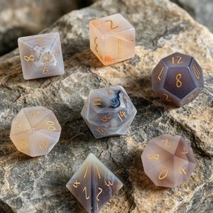 May include: A set of seven polyhedral dice crafted from polished agate stone in shades of gray, beige, and translucent white. Each die features engraved numbers filled with gold-colored ink, showcasing natural mineral banding and inclusions.