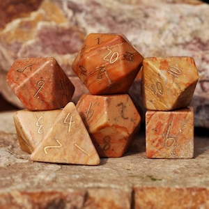May include: A set of seven stone dice in shades of orange and tan, with gold-colored numbers. The dice include a tetrahedron, a cube, a dodecahedron, and a 20-sided die. The dice are arranged on a stone surface.