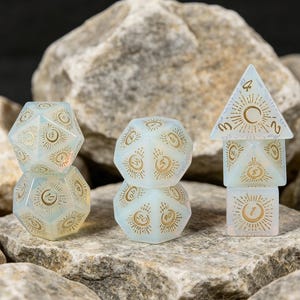 May include: A set of polyhedral dice made of translucent, milky white material with gold-coloured engravings. Each face features a sunburst design surrounding a central crescent moon and a number. The dice are stacked in three columns on a rocky surface.