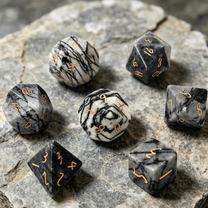 May include: A set of seven polyhedral dice, each with a unique shape and number of sides. The dice are made of a gray stone with black veining and gold-colored numbers. The dice are arranged on a gray stone surface.