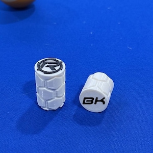May include: Two white cylindrical objects with black logos and text, possibly for a pool cue. One features a stylized "G" logo, the other reads "BK". They have a textured, geometric pattern. An eight ball is in the background.