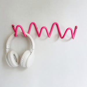 Könnte beinhalten: Eine leuchtend pinke Wandgarderobe mit einem durchgehenden wellenförmigen Zickzack-Design und mehreren Haken. Ein weißer Over-Ear-Kopfhörer hängt am linken Haken der Metallgarderobe, die mit Schrauben an der Wand befestigt ist.