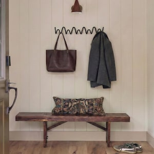 Könnte beinhalten: Ein rustikaler Eingangsbereich mit einer Holzbank, einer dunkelbraunen Ledertasche und einem grauen Mantel, der an einer schwarzen Metallgarderobe hängt. Ein dekoratives Kissen liegt auf der Bank und ein Paar Turnschuhe steht auf dem Boden.