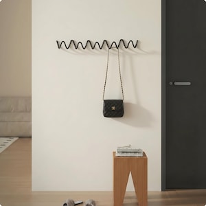 May include: A black, wavy metal coat rack mounted on a white wall, holding a black quilted purse with a gold chain strap. A small wooden stool with books sits below. Slippers rest on the floor.