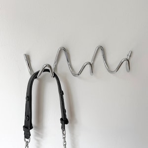Könnte beinhalten: Ein moderner Wandhaken aus Metall in Silber mit wellenförmigem Design. Eine schwarze Lederhandtasche mit silberner Kette hängt an dem Haken. Der Haken ist an einer weißen Wand befestigt.