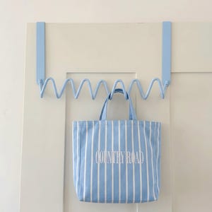 May include: A light blue over-the-door organizer with a wavy design, holding a blue and white striped tote bag. The tote bag has the words "COUNTRY ROAD" printed on it.
