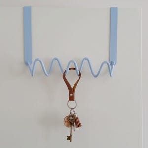 Könnte beinhalten: Ein hellblaues Schlüsselregal für die Tür mit einem wellenförmigen Design. Ein brauner Leder-Schlüsselanhänger mit Schlüsseln und einer Quaste hängt an dem Regal. Die Schlüssel umfassen einen goldfarbenen Schlüssel und ein rundes Etikett mit "HOME".