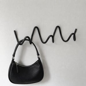 Puede incluir: Un perchero de pared negro con un diseño metálico continuo, ondulado y serpenteante con múltiples ganchos. Un bolso de mano pequeño de cuero negro en forma de media luna con una cremallera de tono plateado cuelga de uno de los ganchos.