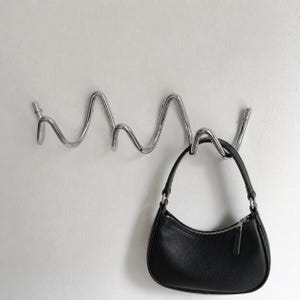 Op de afbeelding: Een golvende, verchroomde metalen wandhaak met een zwarte handtas eraan. De haak is aan een witte muur bevestigd. De handtas heeft een ritssluiting en een korte handgreep.
