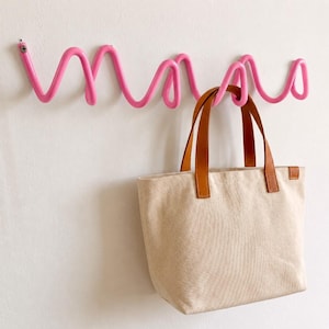 Könnte beinhalten: Eine leuchtend pinke Wandgarderobe mit einem durchgehenden, wellenförmigen Design. Die Garderobe ist mit sichtbaren Schrauben an der Wand befestigt und trägt eine beigefarbene Canvas-Tasche mit zwei braunen Ledergriffen, die an einer Schlaufe hängt.