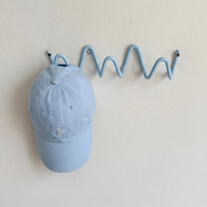 Peut inclure: Une casquette de baseball bleu clair est suspendue à un crochet mural ondulé en métal de la même couleur. La casquette a une visière incurvée et un petit logo blanc brodé. Le crochet est fixé sur un mur blanc.