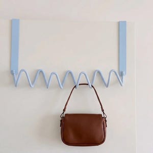 Op de afbeelding: Een lichtblauw over-de-deur haakrek met een golvend ontwerp, met een bruine leren handtas. Het rek is gemonteerd op een witte deur en biedt een ruimtebesparende opbergoplossing voor accessoires.