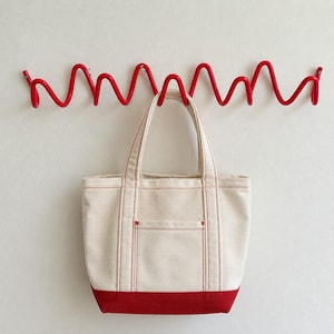 Op de afbeelding: Een crèmekleurige canvas tas met rode stiksels en een effen rode onderkant, hangend aan een felrode, horizontale, golvende metalen wandkapstok met meerdere haken voor opslag en organisatie.