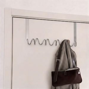Könnte beinhalten: Ein silberfarbener Türhaken mit wellenförmigem Design, an dem eine braune Handtasche und ein grau-weiß gemusterter Mantel hängen. Die Garderobe hat mehrere Haken zum Aufhängen von Gegenständen.