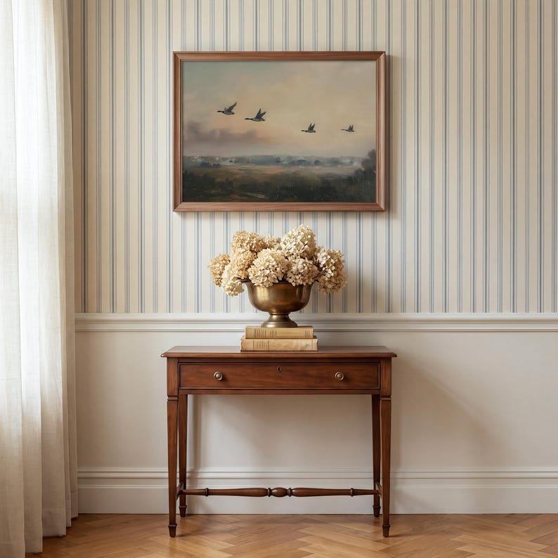 Puede incluir: Un cuadro de paisaje rectangular enmarcado que muestra cuatro aves en vuelo sobre un horizonte de tonos apagados, colocado sobre una mesa consola de madera. Un cuenco de lat&oacute;n con hortensias secas descansa sobre dos libros apilados.