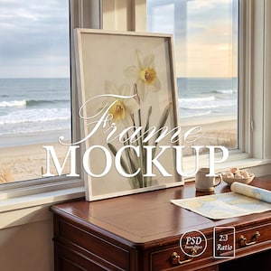 May include: A framed floral print featuring white and yellow daffodils. The frame is white and rests on a dark wooden desk near a window overlooking a beach. The image includes the text "Frame MOCKUP".