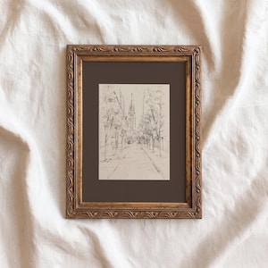 May include: A framed sketch of a city street scene. The artwork, in black and white, depicts a tree-lined road leading towards buildings with tall spires. The frame is ornate, with a gold finish, and the artwork is set against a dark brown mat.