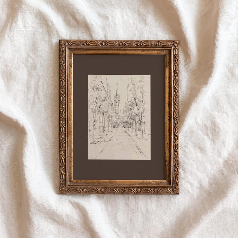 May include: A framed sketch of a city street scene. The artwork, in black and white, depicts a tree-lined road leading towards buildings with tall spires. The frame is ornate, with a gold finish, and the artwork is set against a dark brown mat.