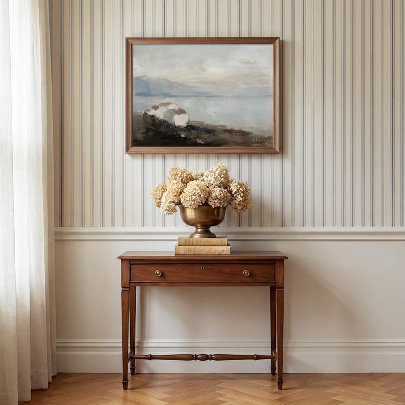 Puede incluir: Un cuadro de paisaje en un marco marr&oacute;n cuelga sobre una mesa consola de madera. Sobre la mesa hay un cuenco de lat&oacute;n con pedestal lleno de hortensias secas, apoyado sobre dos libros apilados. La escena se sit&uacute;a frente a un papel pintado a rayas.