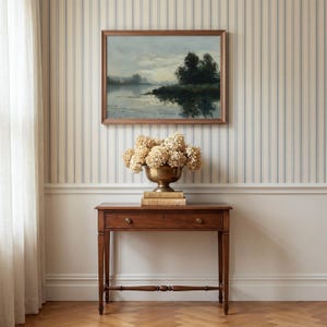 Puede incluir: Un cuadro de paisaje rectangular en un marco de madera marr&oacute;n cuelga sobre una mesa consola de madera marr&oacute;n. Un cuenco de lat&oacute;n lleno de hortensias beige secas descansa sobre dos libros apilados en la mesa, que tiene un caj&oacute;n.
