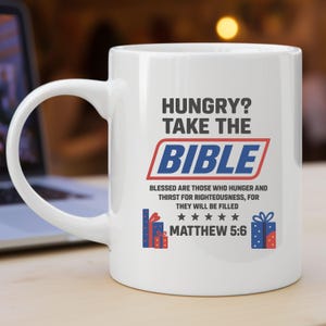 May include: White ceramic mug with the text "HUNGRY? TAKE THE BIBLE" in black and blue. The mug also features the text "BLESSED ARE THOSE WHO HUNGER AND THIRST FOR RIGHTEOUSNESS, FOR THEY WILL BE FILLED" and "MATTHEW 5:6".