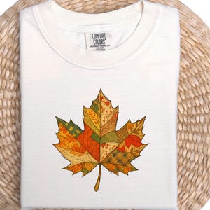 Op de afbeelding: Crèmekleurig T-shirt met een kleurrijk esdoornbladontwerp. Het blad bestaat uit verschillende patchworkpatronen in oranje, geel, groen en bruin. Het shirt is opgevouwen en heeft een label met "COMFORT COLORS".