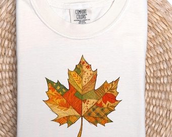 May include: Cream-colored t-shirt featuring a colorful maple leaf design. The leaf is composed of various patchwork patterns in shades of orange, yellow, green, and brown. The shirt is folded and has a tag that says "COMFORT COLORS".