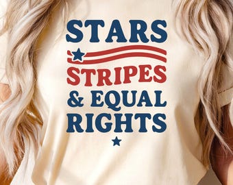 May include: Cream-colored t-shirt featuring the text "STARS, STRIPES & EQUAL RIGHTS" in navy blue and red. The design includes a star and stripes, evoking a patriotic theme. The shirt is a casual wear item.