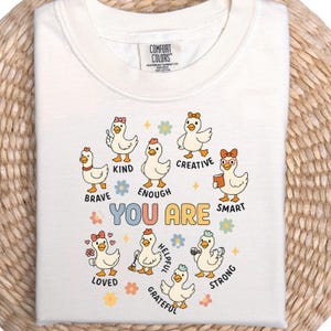 May include: Cream-colored t-shirt featuring a graphic of cartoon ducks with various accessories and the words "YOU ARE" above the words "BRAVE, KIND, ENOUGH, CREATIVE, SMART, LOVED, HELPFUL, GRATEFUL, STRONG". The shirt is made of heavyweight cotton.