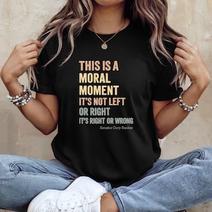 Op de afbeelding: Zwart T-shirt met de tekst "THIS IS A MORAL MOMENT IT'S NOT LEFT OR RIGHT IT'S RIGHT OR WRONG" in een verloop van oranje en geel. De tekst wordt toegeschreven aan senator Cory Booker. Het shirt wordt door een persoon gedragen.