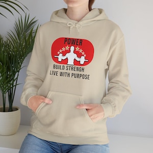 K&ouml;nnte beinhalten: Beigefarbener Kapuzenpullover mit Fronttasche und Kordelzug. Auf der Vorderseite befindet sich ein rotes ovales Grafikdesign mit dem Wort "POWER", einer Gewichtheber-Silhouette und dem Text "BUILD STRENGH LIVE WITH PURPOSE".