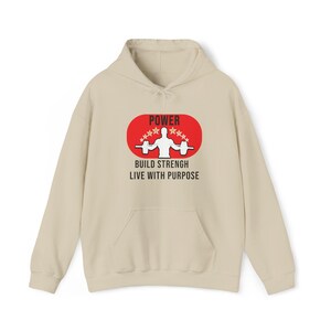 K&ouml;nnte beinhalten: Ein beigefarbener Kapuzenpullover mit einer Vordertasche und einer Kapuze mit Kordelzug. Auf der Vorderseite befindet sich ein rotes Oval mit dem Wort "POWER" &uuml;ber einer Gewichtheber-Grafik und dem Text "BUILD STRENGTH LIVE WITH PURPOSE".