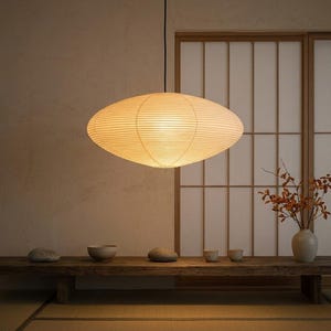 Pode incluir: Uma luminária pendente de formato oval com uma cúpula de papel canelado em tom branco sujo que emite um brilho quente e difuso. A luz pende de um fio preto fino sobre um banco de madeira longo e baixo com uma superfície natural.